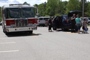 A fire truck and police vehicle are parked in a lot. Several people, including police officers, are gathered around the vehicles. A man and two children stand nearby. Trees and other parked cars are visible in the background. It appears to be a sunny day.