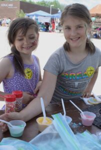 Two smiling young girls with face paint are at an outdoor event. They each wear "I Climbed" stickers and are working with colored powders in cups, likely crafting or making treats at a table. A building and other people can be seen in the background.