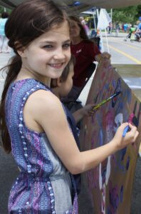 A young girl with long brown hair and a patterned blue dress stands outdoors, smiling as she paints on a large canvas with other children. The scene is lively with people in the background, suggesting a community event or fair.