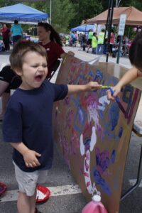 A young boy wearing a dark blue shirt yells playfully while painting on a large canvas at an outdoor event. The canvas is covered with colorful paint marks. Adults and other children are visible in the background under tents.