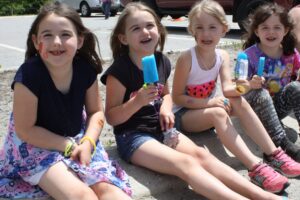 Four young girls sit on a curb in a parking lot, smiling and enjoying popsicles. They have face paint and temporary tattoos. One wears a dress, and the others wear shorts and shirts. A red SUV is parked in the background. It's a sunny day.