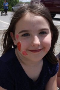 A young girl with long brown hair and a dark blue shirt smiles at the camera. She has face paint on her cheek, including a red heart and other small shapes. The background shows a parked car and an adult figure in the distance.