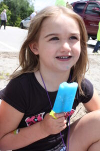 A young child with light brown hair wearing a black shirt is sitting outdoors and holding a blue popsicle. There is a slight blue stain on their chin from the popsicle. A maroon vehicle and a few people are visible in the background.