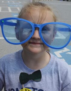 A young child with light hair stands outside on a sunny day, wearing oversized, blue plastic glasses and a large cutout bow tie on their shirt. There is a bit of chocolate or dirt on their cheek and chin. They have a playful and happy expression.