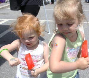 Two young children eating red popsicles stand side by side. The child on the left has curly red hair and is looking at their hand, which has a temporary tattoo. The child on the right has light brown hair, a temporary tattoo of a lollipop on their arm, and looks downwards.