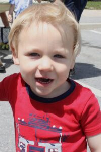A young child with light blonde hair smiles at the camera. The child is wearing a red shirt with a blue and white graphic design. The background includes a blurry outdoor setting with people and pavement.
