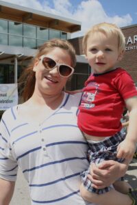 A woman wearing sunglasses and a striped shirt holds a young child in her arms. The child is wearing a red T-shirt and checkered shorts. They are outdoors in front of a building with a banner that reads "Annual Celebration." The sky is clear and blue.
