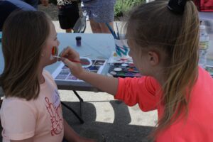 Two girls are at a face painting station outdoors. One girl in a pink shirt sits patiently while the other girl in a bright pink top paints a rainbow on her cheek. The background shows a table with painting supplies and people standing around.