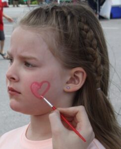A young girl with braided hair has a concerned expression as someone paints a red heart on her cheek. She is wearing a light-colored shirt and there are people in the background.