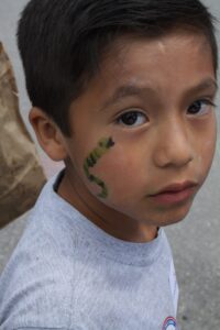 A young boy with short dark hair looks at the camera with a slight expression. He has a green snake face painting on his right cheek and is wearing a light gray T-shirt. The background is blurry, showing a paved surface and part of a paper bag.