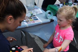 A young girl with blonde hair wearing a pink shirt sits as an artist paints her arm at an outdoor event. The table is covered with painting supplies, water bottles, and pictures of design options. Another individual works at the neighboring table.