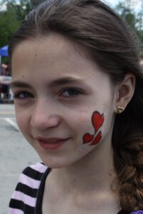 A young girl with long, dark hair and a striped shirt smiles slightly. She has a red and black face painting of two joined hearts on her left cheek, accented by white stars. She is wearing gold heart-shaped earrings, and the background appears to be an outdoor setting.