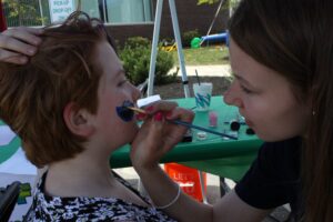 A person with short, red hair sits patiently as a woman applies face paint to their cheek. They are outdoors, possibly at an event or fair. The face paint is blue, and the woman focuses intently on her work while holding a brush and palette.
