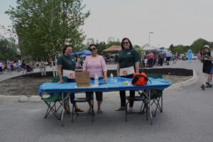 Three individuals stand behind a table at an outdoor event. The table is covered with a blue cloth and holds various items, including water bottles, a red bag, and a cardboard box. Many people and tents are visible in the background, indicating a lively gathering.