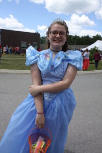 A young girl dressed as Cinderella is standing outdoors at an event under a partly cloudy sky. She is wearing a light blue gown with puffy sleeves, a tiara, and glasses, and is holding an orange bucket filled with what appears to be glow sticks. People and tents are visible in the background.