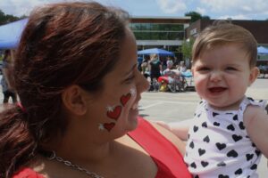A woman with red hearts painted on her cheek holds a smiling baby in her arms. The baby is wearing a white outfit with black hearts. They are outdoors on a sunny day, with people and umbrellas in the background.