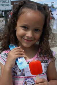 A young girl with curly hair smiles while holding a red ice pop in one hand and dental hygiene items in her other hand. She wears a striped shirt and has a tag hanging around her neck. A parking sign and other people can be seen in the background.
