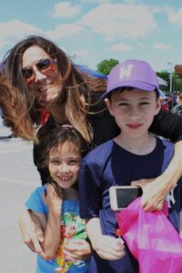 A woman with sunglasses and long hair embraces two children outdoors on a sunny day. The girl in a colorful shirt smiles, holding candy, while the boy in a purple hat and blue shirt holds a pink bag and an electronic device.