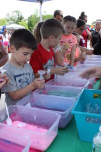 Children gather around a table filled with colorful bins of sand at an outdoor craft event. They use plastic cups and scoops to fill their own containers with different colors of sand. Adults and more children are in the background, engaging in the activity.