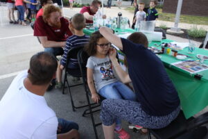 A young girl with curly hair sits on a chair while an adult paints her face at a booth. There are other people around, including children standing and adults seated at a table covered with art supplies. The scene appears to be outdoors at an event.