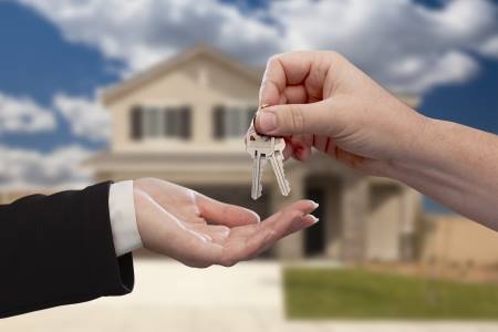 One hand is giving a set of house keys to an open hand in front of a blurred background depicting a house. The image symbolizes the transfer or purchase of a home.