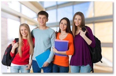 Four smiling young adults, two men and two women, stand close together indoors, holding folders and textbooks. They are casually dressed and wearing backpacks. The background shows a modern, brightly lit building with large windows.
