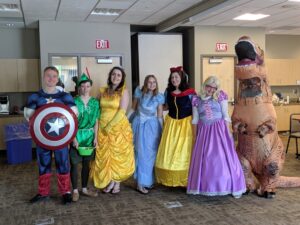 A group of seven people are in a room dressed in various costumes. They are wearing outfits of Captain America, Peter Pan, Belle, Cinderella, Snow White, Rapunzel, and an inflatable T-Rex. The room has beige walls, a screen, and two exit signs above closed doors.