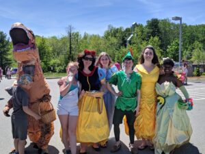 A group of six people pose outdoors in a parking lot, dressed in various costumes including a dinosaur, Snow White, Peter Pan, Belle from Beauty and the Beast, and Tiana from The Princess and the Frog. It's a sunny day with trees in the background.