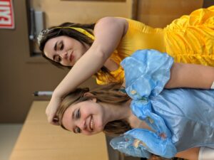 Two women are dressed in elaborate, colorful costumes resembling classic fairytale characters. One in a yellow dress adjusts the hair of the other in a blue dress. Both are smiling in a well-lit indoor setting. An exit sign is visible in the background.
