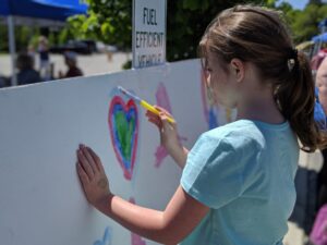 A young girl painting a heart shape on a large white canvas outdoors. She is holding a yellow paintbrush and has a sticker on her hand. The background includes trees and a sign indicating "Fuel Efficient Vehicle." Other people and tents are blurred in the distance.