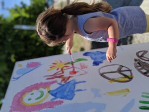 A young child with brown hair in a ponytail is painting on a large outdoor easel. The painting features colorful elements like a smiling figure, peace sign, and various shapes. The child wears a sleeveless gray top and a pink wristband. Trees are in the background.