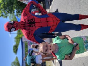 A young girl smiles at the camera while standing next to a person dressed in a Spider-Man costume. The event appears to be outdoors with booths and people in the background. The girl is wearing a green shirt and holds a small toy. The weather is sunny with a clear sky.