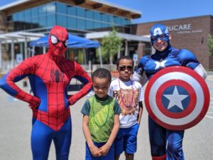 Two children stand between two adults dressed as superheroes: one as Spider-Man and the other as Captain America. They are outside a building with a sign that reads "Bucare Central Maine" in the background. The weather appears to be sunny.