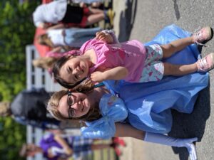 A smiling young woman dressed as a princess in a blue gown and tiara is kneeling next to a smiling small child wearing a pink shirt with a flamingo and floral-patterned shorts. They are outdoors at a lively event with other people and children in the background.