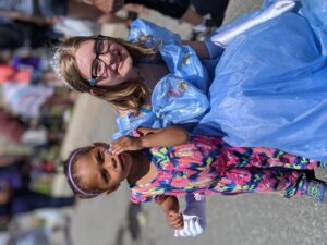 A young girl dressed in a colorful jumpsuit stands beside an older girl wearing a blue princess gown and tiara. They are outdoors with a crowd in the background. The younger girl holds a small object while the older girl smiles at the camera.