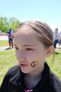 A child with a face painting of a peace sign in red, white, and blue on their cheek looks slightly to the side. The background shows a sunny outdoor setting with a person in a Captain America costume and other people in casual attire.