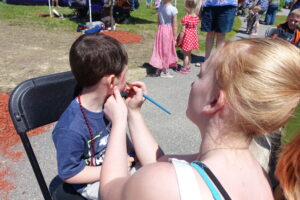 A young boy sits on a chair outdoors, getting his face painted by a woman with light hair. The boy is wearing a dark blue shirt and has a red beaded necklace. Other children and adults are visible in the background, some standing and some walking around.
