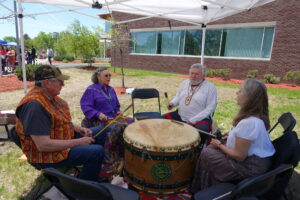 Four people are sitting in a circle under a white canopy, playing a large drum together. Two men and two women, dressed in colorful, traditional attire, are engaged in drumming. There is a brick building and some greenery in the background.