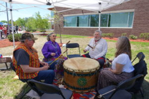 Four people sit in a circle under a canopy, playing a large drum. Two of them are wearing colorful, traditional garments. They are outside, near a brick building and greenery. The group seems to be engaged in a cultural or musical performance.