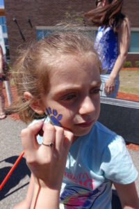 A young girl with light brown hair is having her face painted with a blue paw print on her cheek. She is wearing a light blue shirt and is outside on a sunny day. A woman in the background has long, dark hair and is wearing a blue shirt.