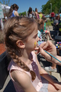 A young girl with brown hair in two braids getting her face painted with a yellow design at an outdoor event. A person's hand is holding a paintbrush, applying the paint. Other people and event activities can be seen in the background under a sunny blue sky.