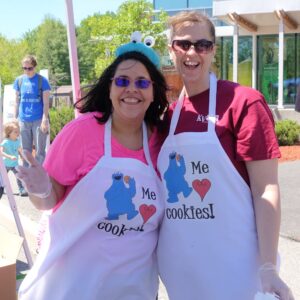 Two women are smiling and posing together outdoors, both wearing white aprons with a Cookie Monster print that says "Me love cookies!" One woman is in a pink shirt and wearing a Cookie Monster headband, while the other is in a maroon shirt. They appear to be at a fun event.