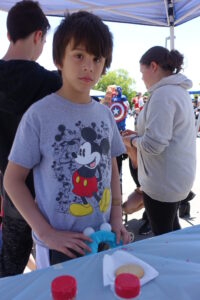 A child wearing a gray Mickey Mouse T-shirt stands under a canopy at an outdoor event. They are standing near a table with a plate of cookies and containers. Other people in casual clothing are nearby, and one person is holding a Captain America shield.