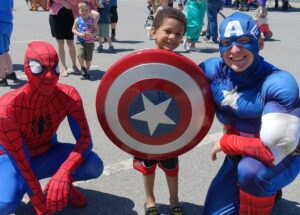A young boy stands outdoors holding Captain America's shield, flanked by two adults dressed in Spider-Man and Captain America costumes. They are all smiling and posing for the camera. In the background, other people are visible and children are playing.