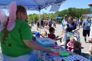 People gather at an outdoor event under blue canopies. A woman with pink fairy wings and a green shirt facilitates bubble-making activities with large wands and bubble trays. Children are engaged and surrounded by adults, strollers, and sunny weather.