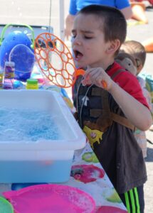 A young child blows bubbles through a large bubble wand at an outdoor event. The child is wearing a backpack with a stuffed monkey attached and is using a soapy solution in a plastic tub. The event appears to have various colorful activities for children.