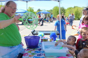 A woman in a green shirt and glasses demonstrates how to make bubbles using a large bubble wand at an outdoor event. Several children gather around a table, eagerly participating. The table is covered with a colorful tablecloth and has a container of soapy water.