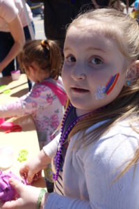 A young girl with a rainbow and cloud painted on her cheek looks towards the camera while participating in an outdoor craft activity. She is wearing a white sweater, beaded necklaces, and is surrounded by craft supplies. Another child is in the background.