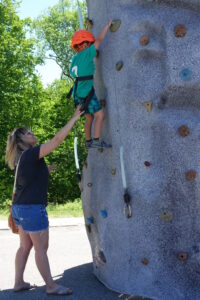 An adult wearing sunglasses and a black T-shirt assists a child in climbing an outdoor rock-climbing wall. The child, donning a bright orange helmet, green shirt, and harness, reaches for a higher hold. Green trees and blue sky are visible in the background.