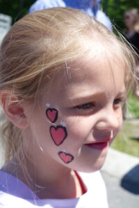 A young girl with face paint featuring three red hearts with black outlines on her cheek is outdoors, smiling slightly. She has blonde hair tied back and is wearing a white shirt. The background is blurred with greenery and people in the distance.